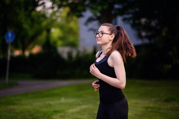 A young woman in black sportswear and glasses jogging in a park, showcasing determination and a healthy lifestyle.