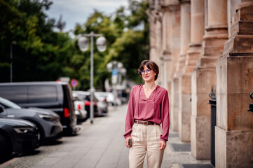 Woman in casual attire smiling while walking on a city sidewalk lined with parked cars and buildings.