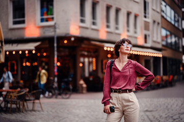 Woman in casual attire stands confidently with her hand on her hip, holding her phone, in a bustling city street with cafes and lights.