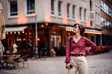 Obraz premium Woman in casual attire stands confidently with her hand on her hip, holding her phone, in a bustling city street with cafes and lights.