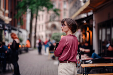 Fototapeta premium Woman in casual attire and glasses, holding her phone and backpack, exploring a bustling city street lined with cafes and people.