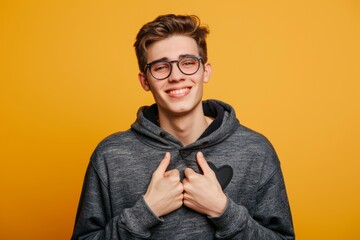 Fototapeta premium A young man wearing glasses and a grey hoodie with a black heart on it smiles and gestures towards his heart while standing in front of a yellow background