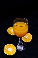 Orange juice in glass and half oranges next to the glass. Orange juice on black background. Background of fresh juice with orange fruit with concept of still life, vegan, healthy fruit, and elegance.