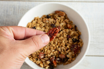 A top down view of a hand holding a pecan over a bowl of granola cereal.