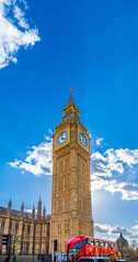 Obraz premium View from below of the Big Ben Clock Tower seen in great detail of its golden ornamentation and a red double-decker London bus traveling on a bright day with clear blue sky and backlit clouds.