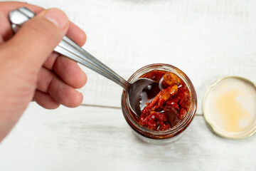 A top down view of a hand using a spoon to scoop up some sun-dried tomatoes from a jar.