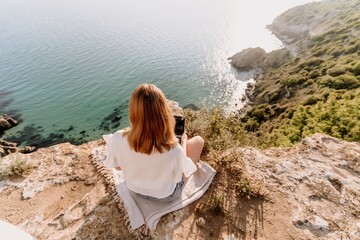 A woman is sitting on a rock overlooking the ocean with a laptop in front of her. She is enjoying the view and the peacefulness of the location.