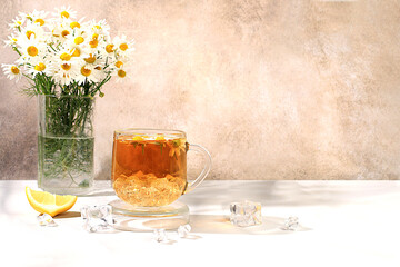Chamomile tea with lemon in a transparent cup with natural small chamomile flowers on a light rustic table, the concept of herbal tea and proper natural nutrition, summer card,