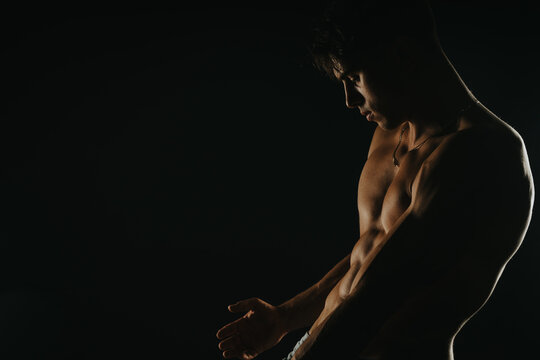 Shirtless muscular young man posing in low light, showcasing his defined physique and strength. Dark and dramatic lighting creates a moody and intense atmosphere.