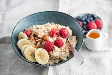 Cooked oatmeal porridge in a bowl served with raspberries, banana slices and chopped almonds. Healthy breakfast choice