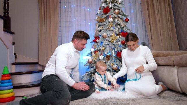 Happy Caucasian family are in the room decorated for Christmas. Parents and their baby play with the string of beads.