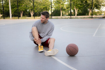young man in athletic wear sits on an outdoor basketball court, holding his ankle in pain after suffering an injury