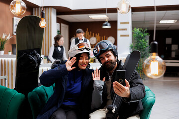 Tourists enjoying winter vacation at ski resort dressed in snow gear. Young couple use mobile device to capture the moment while surrounded by employees and wintersport equipment in hotel lobby.