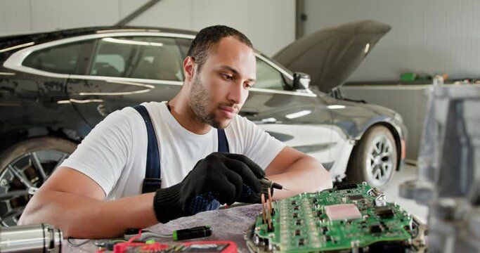 Male Mechanic Repairing Electric Car's Control Unit. Mechanic working on an electric car's control unit in a workshop. The service includes voltmeter usage and intricate engine repairs.