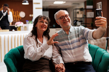 Elderly caucasian couple on vacation utilising mobile phone for video chat in exclusive hotel...