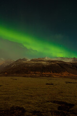 Aurora borealis over countryside scenery, snow covered mountain slopes and glowing sky filled with stars. Northern lights in Iceland creating magical unique landscape, green and black colors.