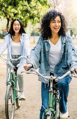 Vertical photo. Two young multiracial women having fun riding bike together in the park. Smiling female friends enjoying time together on summer vacation. Friendship and holidays concept. © Xavier Lorenzo