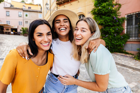 Smiling portrait of small group of three young women laughing at camera standing together at picturesque village during summer vacation. Holiday lifestyle and friendship concept
