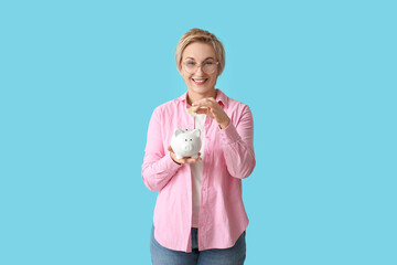 Happy mature woman putting coins into piggy bank on blue background