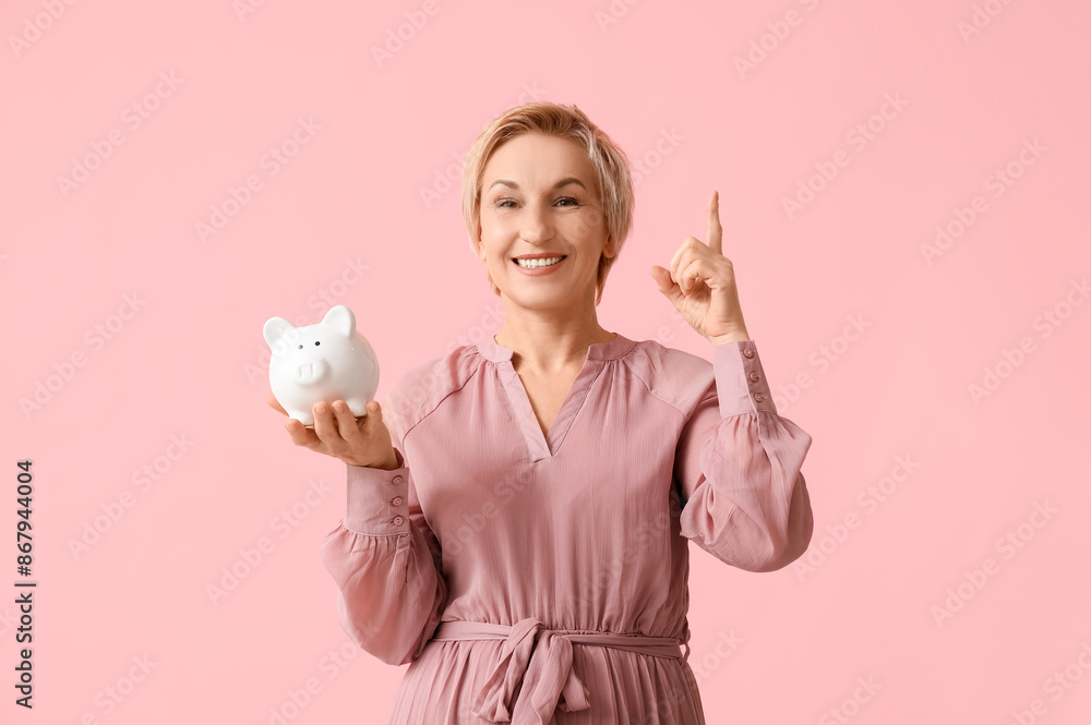 Poster Beautiful mature woman with white piggy bank pointing at something on pink background