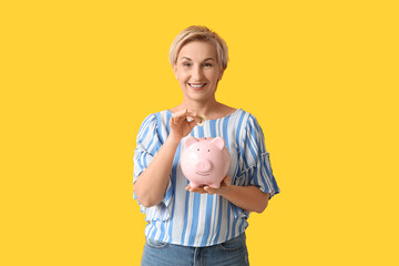 Beautiful mature woman putting coins into pink piggy bank on yellow background