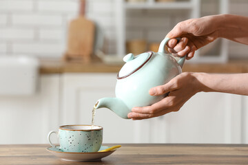 Woman pouring tea from teapot into cup on table in kitchen