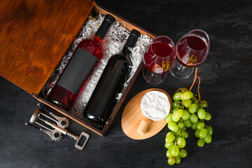 Box with bottles of wine, glasses, grapes and Camembert cheese on black grunge background