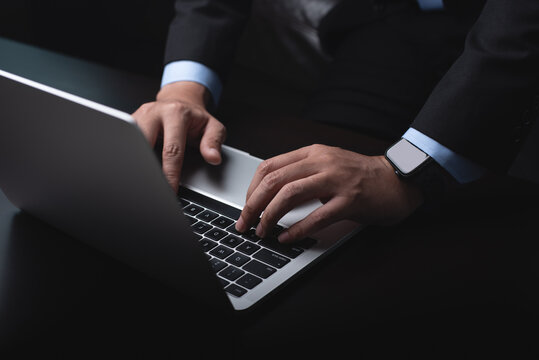 Businessman working on laptop computer, hand typing on keyboard on table at office with dark background, online working, close up