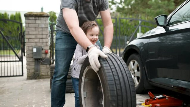Cute toddler boy helping his father to change car wheels in their backyard. Father teaching his little son to use tools. Active parent of a small child. Slow motion summer day footage.
