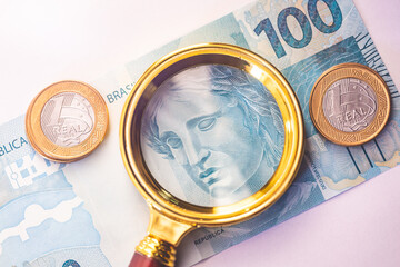 Dinheiro, Real - BRL, moeda do Brasil. Uma nota de 100 reais com uma lupa dourada e duas moedas de...