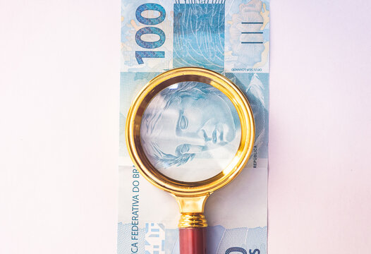 Vista superior de uma lupa dourada sobre uma nota de 100 reais do Real Brasileiro - BRL. Fundo branco. Economia, Brasil, desemprego e infla&ccedil;&atilde;o.