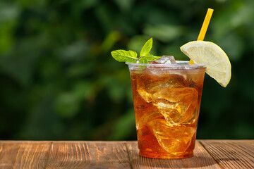 plastic glass with ice tea and lemon on wooden table with green garden on the background