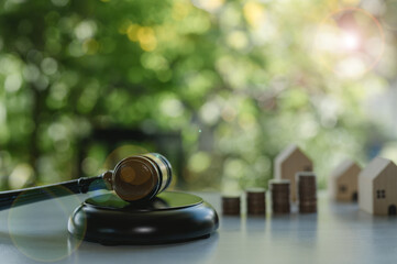 A gavel sits on a wooden surface with a stack of coins and a house model. The scene suggests a courtroom setting, with the gavel symbolizing authority and the coins