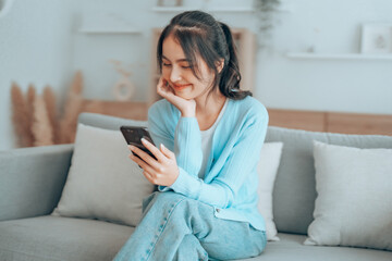 Obraz premium Smiling pretty young Asian woman sitting on cozy couch, using funny mobile apps in living room. Woman at Home, Doing Online Shopping, Messaging Friends, Posting on Social Media