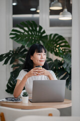 A young woman holding a coffee cup, working on a laptop in a modern home office with indoor plants and natural light.