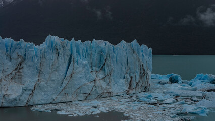 A wall of cracked blue ice with sharp peaks and crevices rises above the glacial lake. Thawed ice...