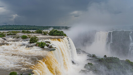 An impressive tropical waterfall landscape. The streams of the stormy river collapse into the abyss. Rocks with green vegetation in a turbulent riverbed. The spray rises like a cloud into the blue sky
