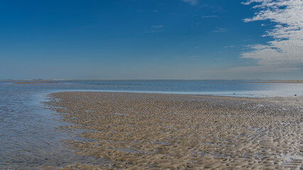 Obraz premium The low tide in the ocean. A bizarre natural pattern of sandy ridges, puddles, and streams formed on the exposed seabed. Waves are foaming on the horizon. Clouds in the blue sky. Copy space. 