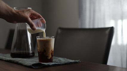 man pours milk into fresh coffee over in glass on wood table