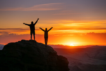 Two silhouettes of happy couple are standing with open arms on big rock against sunset sky and sun