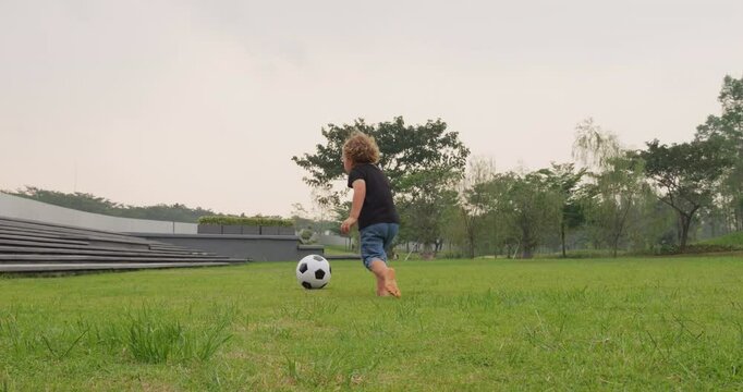 Toddler boy receives light soccer pass but swings too early and misses ball. Unfazed, he joyfully chases rolling ball, catches up, and kicks it towards camera. Relaxed game or simple training at park.