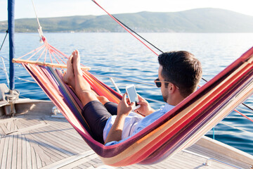 Man with mobile phone relaxing on yacht in hammock. Sea travel on sailboat. Traveler gaming on...