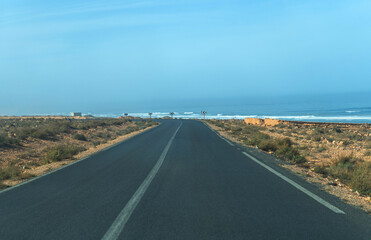Fototapeta premium Road to ocean in Morocco, straight path, arid landscape, sparse vegetation, clear sky, distant waves
