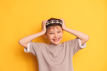 A smiling boy puts a Kazakh skullcap on his head against a yellow background.