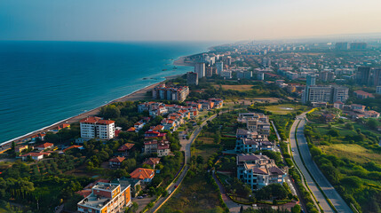 Aerial view of a coastal city with a mix of residential buildings, green spaces, and a vast blue sea under a clear sky.