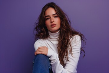 Fototapeta premium A young woman with long wavy hair, wearing a white turtleneck and jeans, sitting cross-legged against a solid purple background, looking relaxed and contemplative.