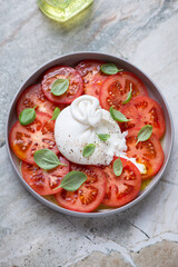 Plate with sliced burrata, red tomatoes and green basil, vertical shot on a grey granite background, elevated view