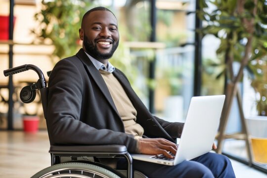 Happy young black man with disability sitting in wheelchair in office Happy young black man with disability sitting in wheelchair in office and looking at camera while analyzing online data on laptop 