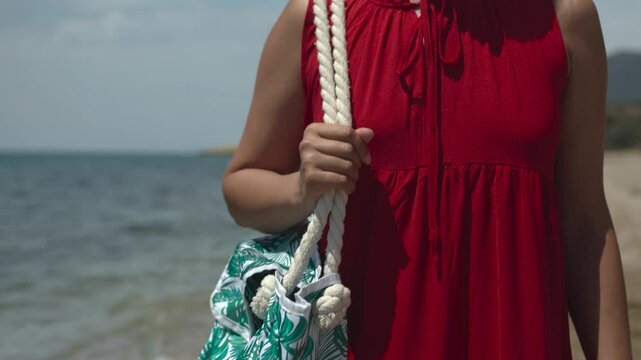 Backwards follow close up shot of young woman wearing a red dress and carrying a beach bag walking along seashore in the summer season  