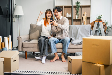  A young couple sits on the sofa, happily taking photos together and inspecting fingernail tabs while packing their belongings into a large box for their move to a new home. They share smiles